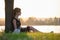 Lonely woman sitting alone on green grass lawn leaning to tree trunk on lake shore on warm evening. Solitude and