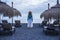 Lonely woman viewed from back side stand in the middle of empty seats and sun umbrellas waiting and looking at the quiet ocean