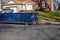 Long blue dumpster on a street by a curb near residential home.