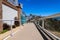 A long concrete footpath along a hillside with a white wooden hand rail with three stone arches covered in red flowers