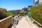 A long concrete footpath along a hillside with a white wooden hand rail with three stone arches covered in red flowers