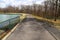 A long curved footpath with a black metal hand rail around a still green lake surrounded by bare winter trees