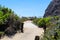 A long dirty footpath to the ocean surrounded by brown wooden posts with rope and lush green trees and plant