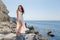 Long-haired girl in wedding dress standing on stone barefoot. Dark-haired young woman in white sleeveless dress posing on coastal