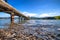 A long pier at Akaroa, New Zealand. The water is so clear that one can see pebbles under the water. There are rolling hills in the