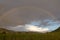 Long rainbow in front of gray rain cloud, green wheat field in the foreground.