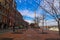 A long red brick sidewalk along the riverfront with rows of bare trees and tall red brick buildings and parked cars