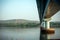 Long road bridge over the river, side view. Powerful stone columns of bridge are reflected in the river water against the shore