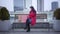 Long shot of cheerful Caucasian girl sitting under light rain on bench in city and talking on the phone. Young pretty