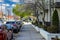 A long sidewalk with apartment buildings, lush green trees and plants and parked cars along the street with blue sky and clouds