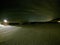 Long snowy field at night with barn and ligh