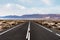 Long straight empty asphalt road through arid landscape against ocean and mountain range