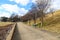 A long straight footpath in the park with bare winter trees, park benches and yellow and green grass along the path with blue sky