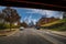 A long street with a yellow line and cars and trucks parked along the street underneath a red metal bridge