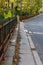 Long view of a very narrow sidewalk bordered by street and rusted wrought iron fencing, trees with leaves