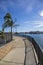 A long winding concrete footpath with a black metal handrail, lush green palm trees, rippling blue water, boats and yachts