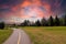 A long winding footpath with a yellow line in the park surrounded by lush green grass and lush green trees with powerful red cloud