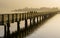 Long wooden boardwalk pier over water in golden evening light with a mountain landscape and people walking silhouette in the backg