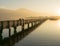 Long wooden boardwalk pier over water in golden evening light with a mountain landscape and people walking silhouette in the backg