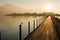 Long wooden boardwalk pier over water in golden evening light with a mountain landscape and people walking silhouette in the backg