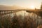 Long wooden boardwalk pier over water in golden evening light with a mountain landscape silhouette in the background and golden ma