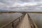 Long wooden pier and boardwalk in brackish water wetlands with esparto grass and lagoon under an overcast sky