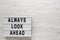 `Always look ahead` words on a lightbox, clipboard with blank sheet of paper on a white wooden surface, top view. Overhead, from