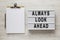 `Always look ahead` words on a lightbox, clipboard with blank sheet of paper on a white wooden surface, top view. Overhead, from