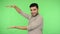 Look here! Happy satisfied brunette man in shirt holding palm and pointing copy space. studio shot, green background,