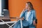 Looking far away. Businesswoman with curly blonde hair indoors in room with orange colored wall and wooden table