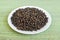 A lot of black pepper grains on a white saucer on a green table mat made of natural plant fibers. Natural food spices and