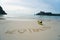 We love Thailand - text written by hand in sand on a sea beach with kayak over sky.