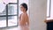 Lovely attractive girl in loose light dress with open bare back, standing at huge window, smiling tenderly, mysterious