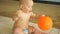 Lovely baby is playing with an orange ball while sitting in a warm children`s pool in an aquapark