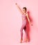 Lovely blond model in tight rose sports clothing dancing on her toes happily, raising one hand up. portrait isolated on pink.