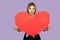 Lovely cute woman holding huge red heart and looking at camera with toothy smile, showing symbol of love and charity