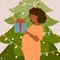 Lovely dark skin pregnant woman is holding Christmas present in her hands near the tree.