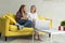 Lovely lesbian couple in casual clothes sitting togethe on yellow sofa with cup of tea