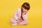 Lovely little girl sitting on floor with crossed legs, keeping palm on chin, looking bored at camera, female child wearing rosy