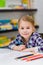 Lovely smiling little girl with blond hair sitting at table with multicolored pencils and looking at camera