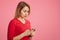 Lovely worried surprised female reads unexpected message on smart phone, wears red sweater, stands sideways against pink backgroun