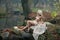 Lovely young lady sitting near river in enchanted woods. Sensual blonde with white clothes posing provocatively in autumnal park.