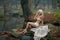 Lovely young lady sitting near river in enchanted woods. Sensual blonde with white clothes posing provocatively in autumnal park.