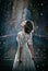 Lovely young lady wearing elegant white dress enjoying the beams of celestial light and snowflakes falling on her face