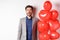 Lovers day. Excited handsome man in suit standing near red hearts balloons, raising eyebrows and looking surprised