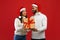 Loving arab couple holding Christmas gift box and smiling to each other, wearing Santa hats, posing over red background