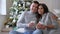 Loving couple with white cups look at camera on couch in morning on background of decorated xmas tree