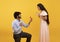 Loving indian man standing on one knee and offering engagement ring to his beloved woman on yellow background