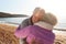 Loving Retired Couple Hugging Standing By Shore On Winter Beach Vacation Against Flaring Sun