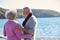 Loving Retired Couple Hugging Standing By Shore On Winter Beach Vacation Against Flaring Sun